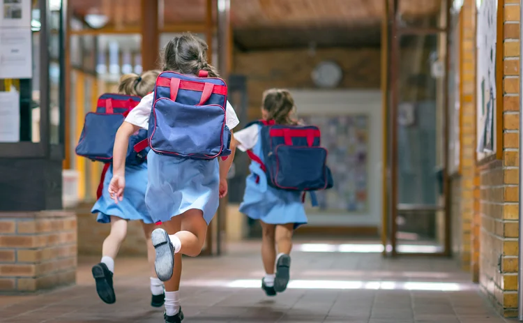 Rear view of excited students running towards entrance. Girls are carrying backpacks while leaving from school. Happy friends are wearing school uniforms.