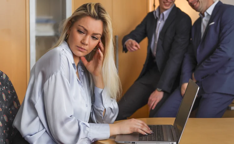 Two men ridicule a woman sitting at her laptop and trying to work.