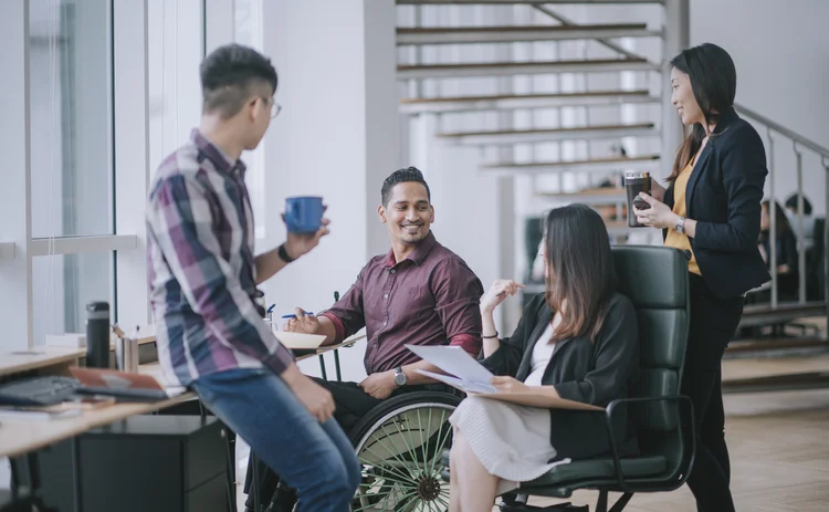 Indian white collar male worker in wheelchair having cheerful discussion leading conversation with colleague in creative office workstation beside window