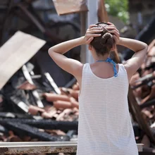 woman standing in front of burned out house and holding her head with both hands